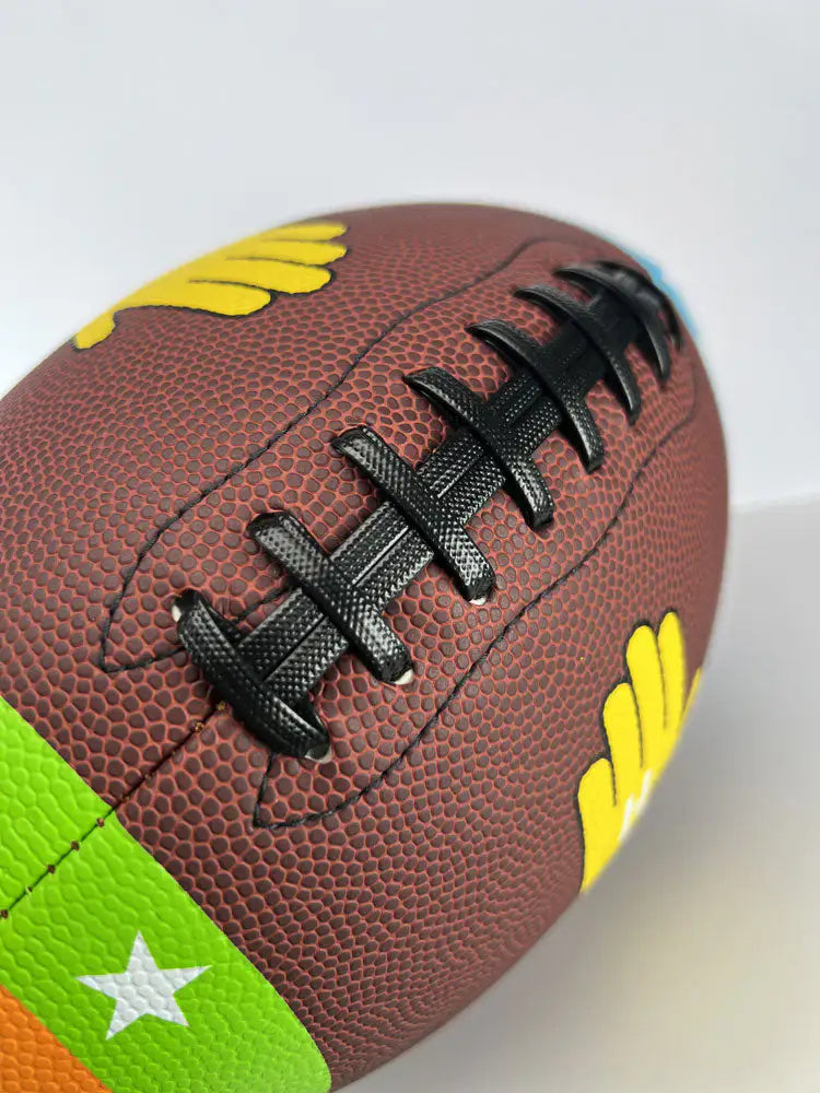 Close-up angled view of the Heisman Factory ColorTech Fundamental Football featuring raised black laces and colorful training panels on a textured brown football.