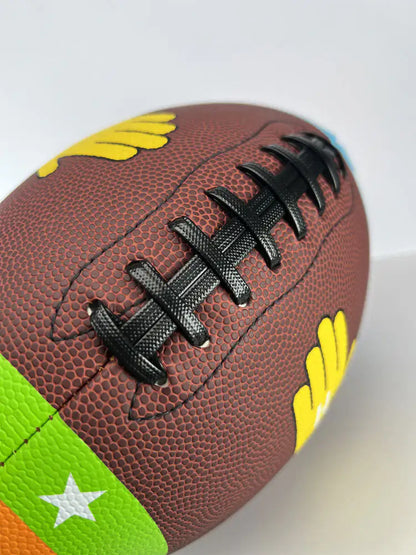Close-up angled view of the Heisman Factory ColorTech Fundamental Football featuring raised black laces and colorful training panels on a textured brown football.